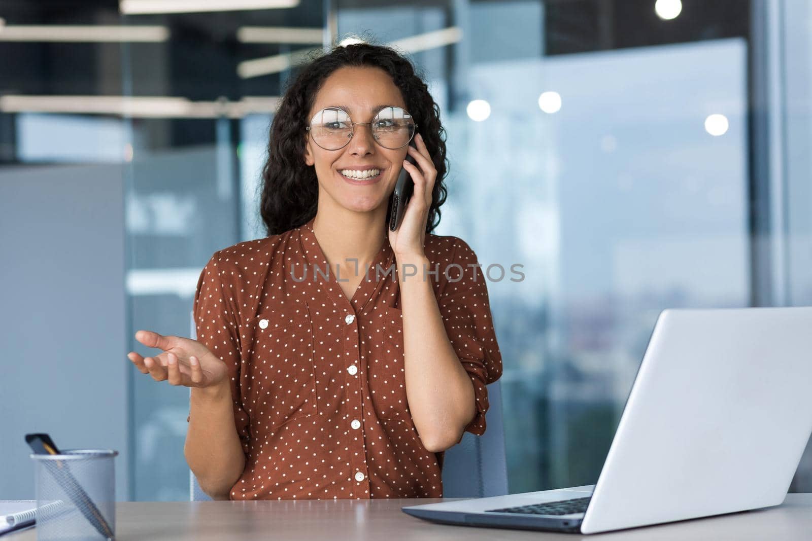 Young hispanic female freelancer wearing glasses sitting in office at desk with white laptop by voronaman