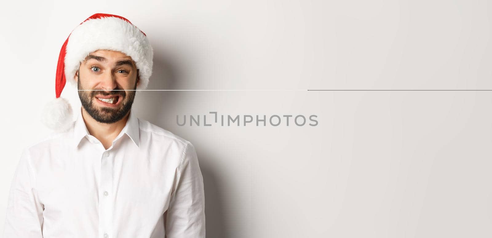Close-up of awkward guy in santa hat apologizing, feeling uncomfortable, standing over white background. Christmas concept by Benzoix
