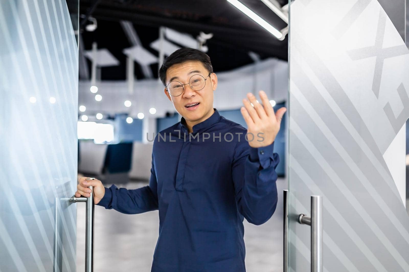 Asian businessman boss invites to his team, man looks at camera and waves, manager in glasses and shirt works inside modern office building.