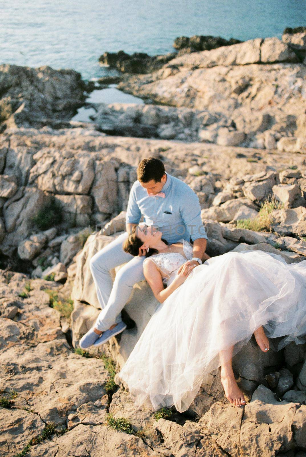 Bride lies on a large stone on the lap of the sitting groom by the sea. High quality photo