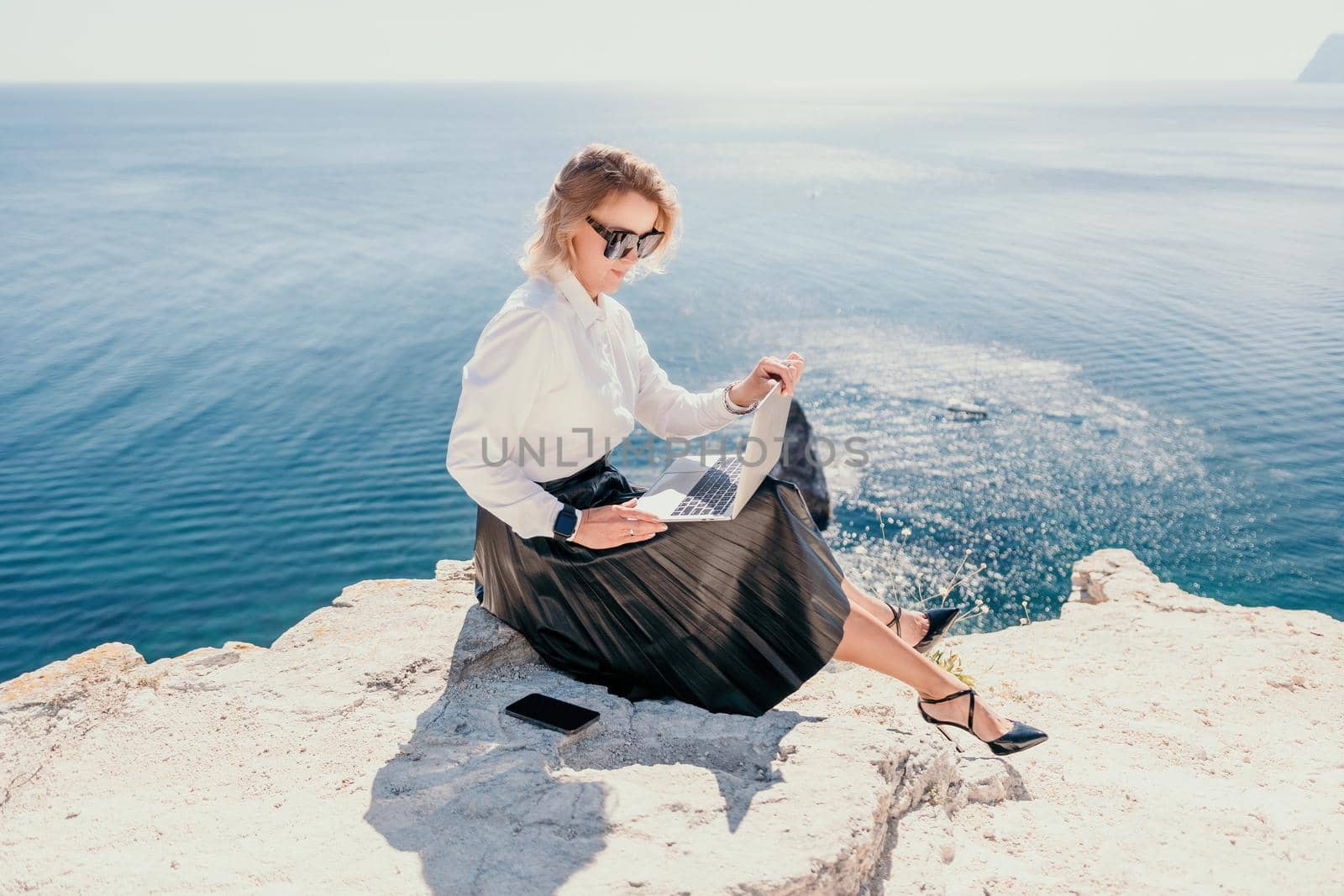 Digital nomad, Business woman working on laptop by the sea. Pretty lady typing on computer by the sea at sunset, makes a business transaction online from a distance. Freelance, remote work on vacation by panophotograph