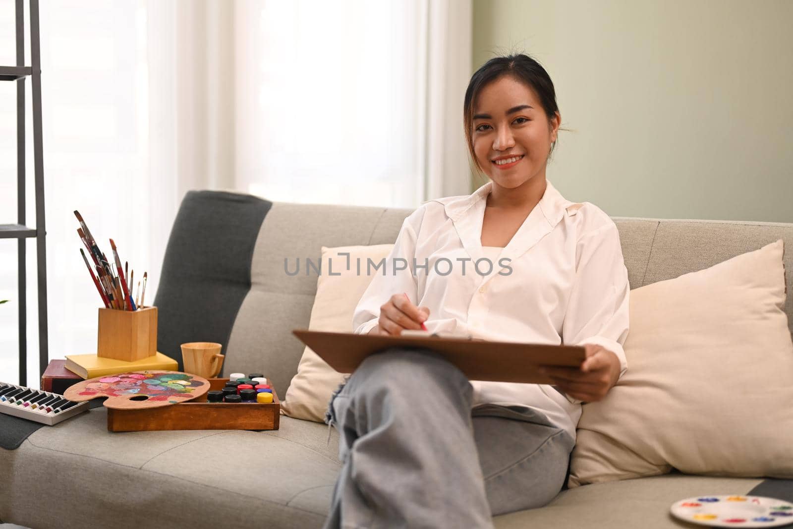 Smiling creative woman painting with watercolor in bright cozy living room. Art, creative hobby and leisure activity concept.