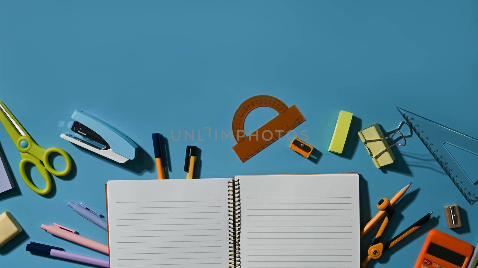 Flat lay blank notepad surrounded by various school supplies on blue background by prathanchorruangsak