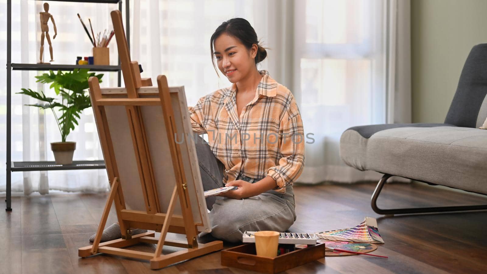 Pleasant female artist sitting on floor in bright living room and painting with watercolor on canvas. Art and leisure activity concept.