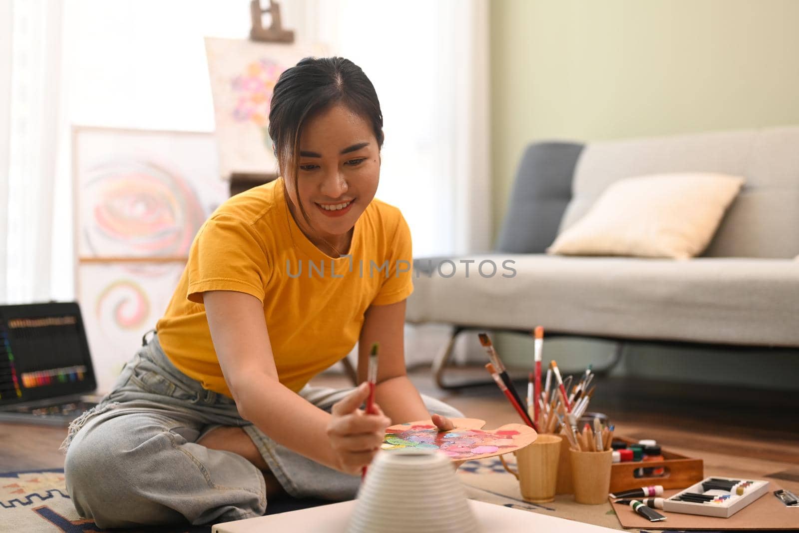 Happy woman painting picture on canvas with oil paints in bright living room. Leisure activity, creative hobby and art concept.