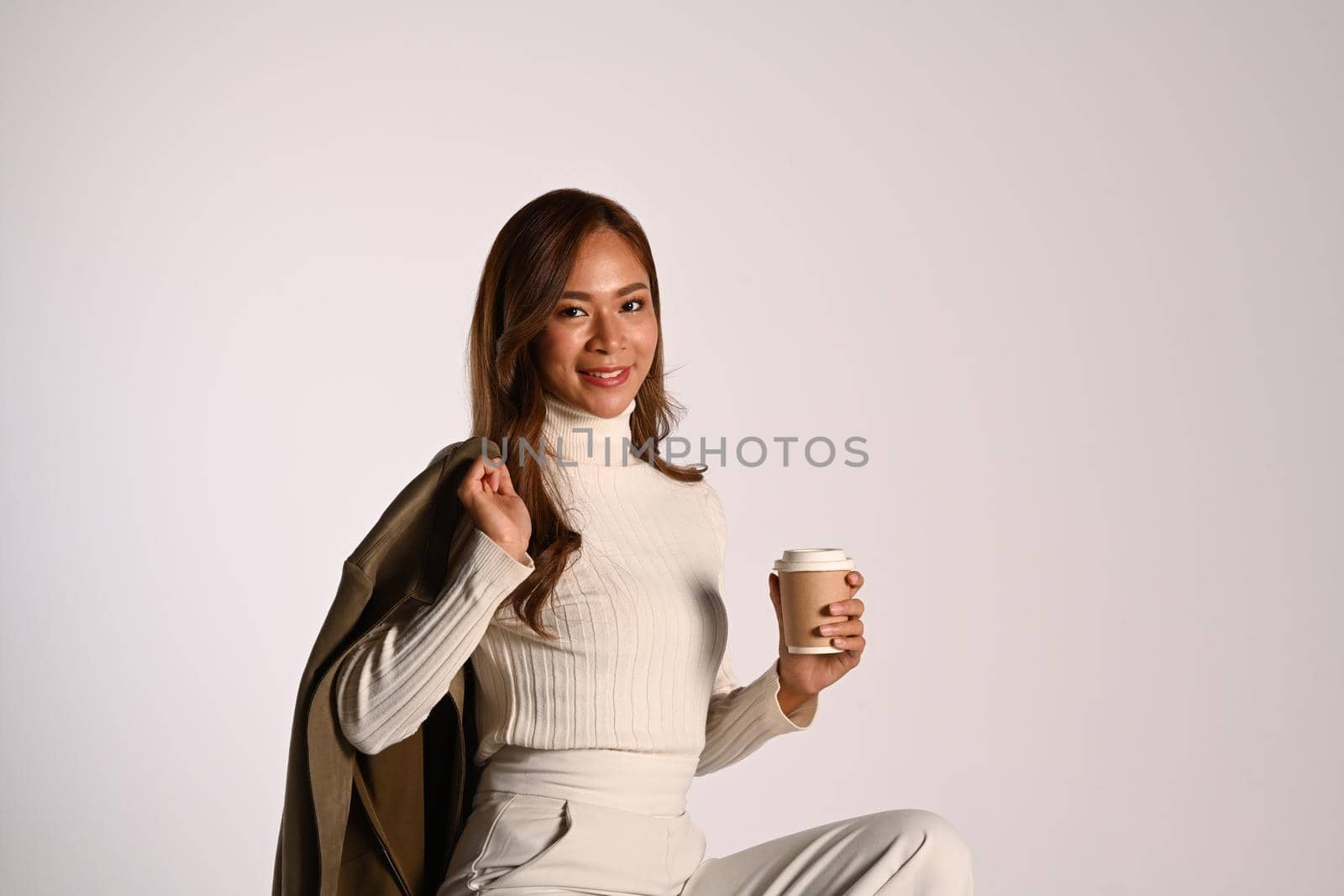 Portrait of stylish young woman in trendy clothes holding paper cup sitting over white background. Fashion, beauty concept.