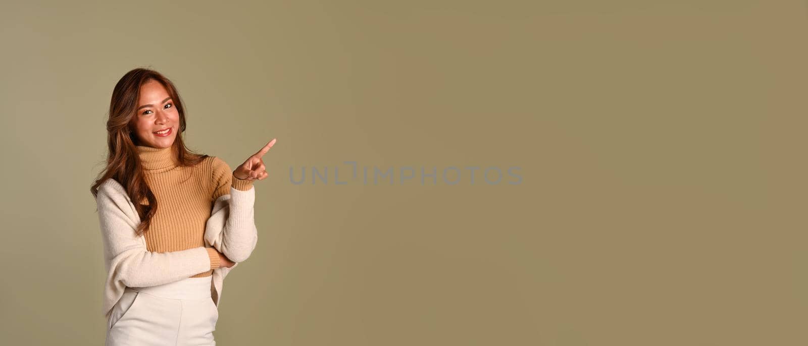 Smiling beautiful woman wearing warm sweater pointing at copy space over beige background. Empty space for your advertise text.