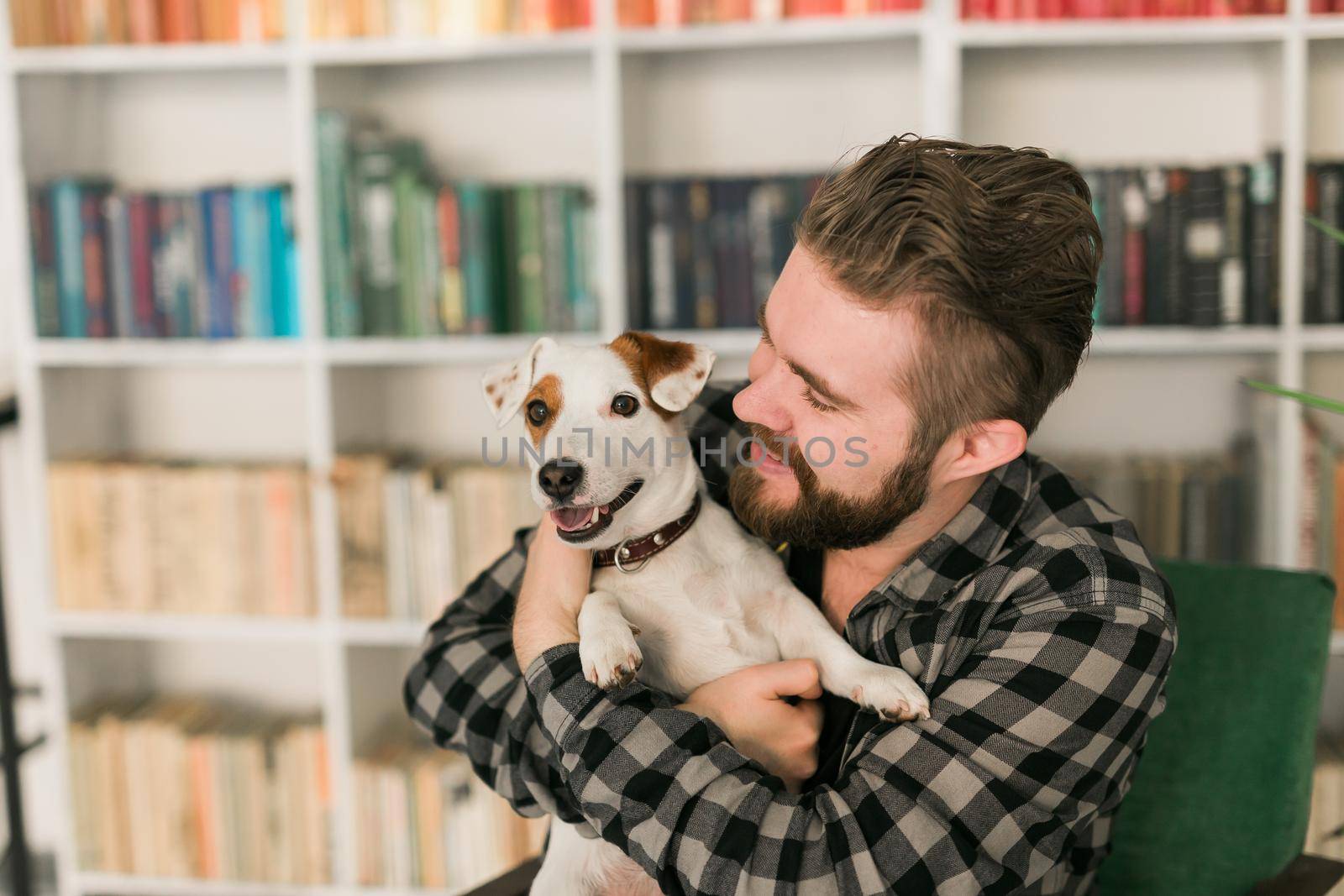 Happy male owner of jack russell terrier dog, feels responsibility of caring about pet, standing against bookshelves background. People and relationship with animals by Satura86