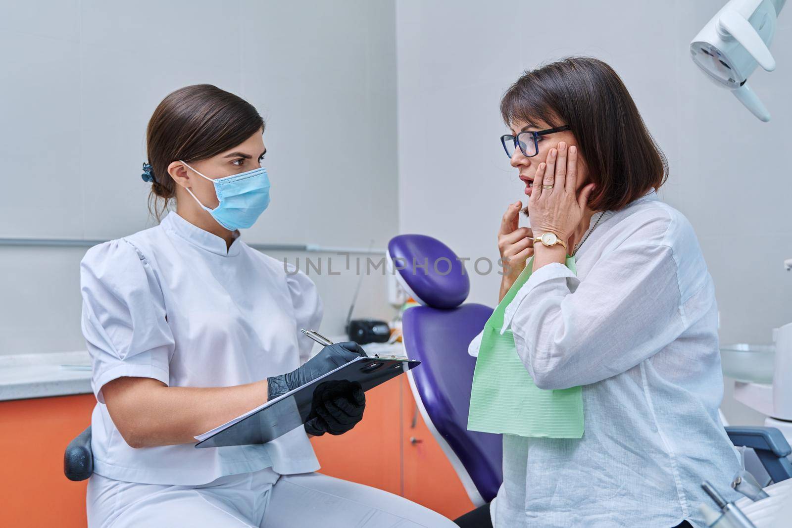 Dentist and woman patient in dental clinic, doctor nurse writing on clipboard consulting, middle aged female talking about toothache disturbing dental problem. Dentistry, treatment, dental health care