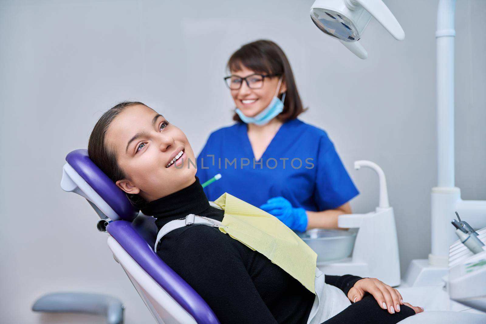 Portrait of young female patient with toothy smile looking at camera sitting in dental chair with dentist doctor. Dentistry, hygiene, treatment, dental health care concept