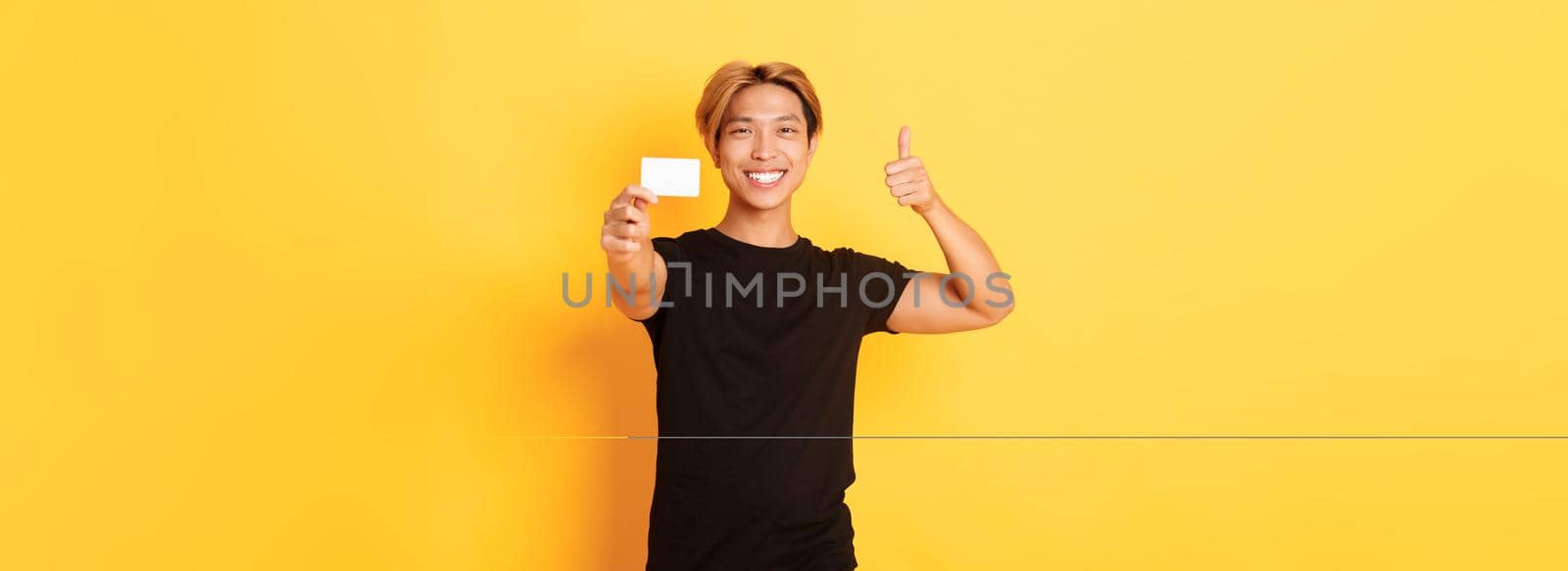 Satisfied handsome asian guy smiling pleased and showing credit card, thumbs-up in approval, standing yellow background.