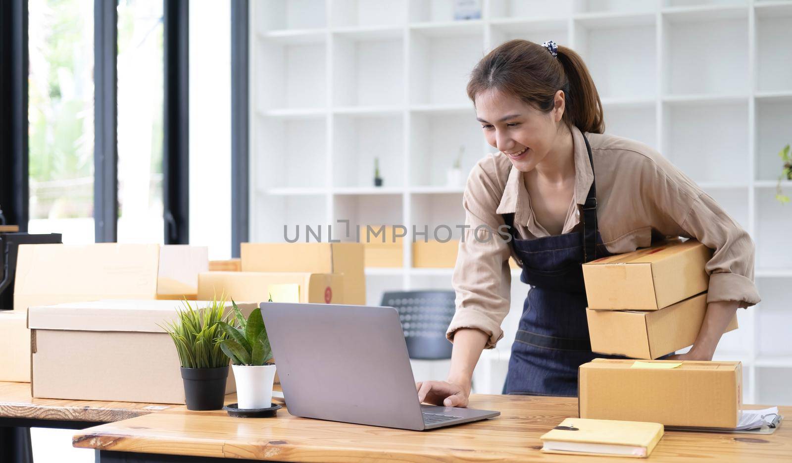 Young smiling beautiful owner asian woman freelancer sme business online shopping working on laptop computer with parcel box at home - SME business online and delivery concept.