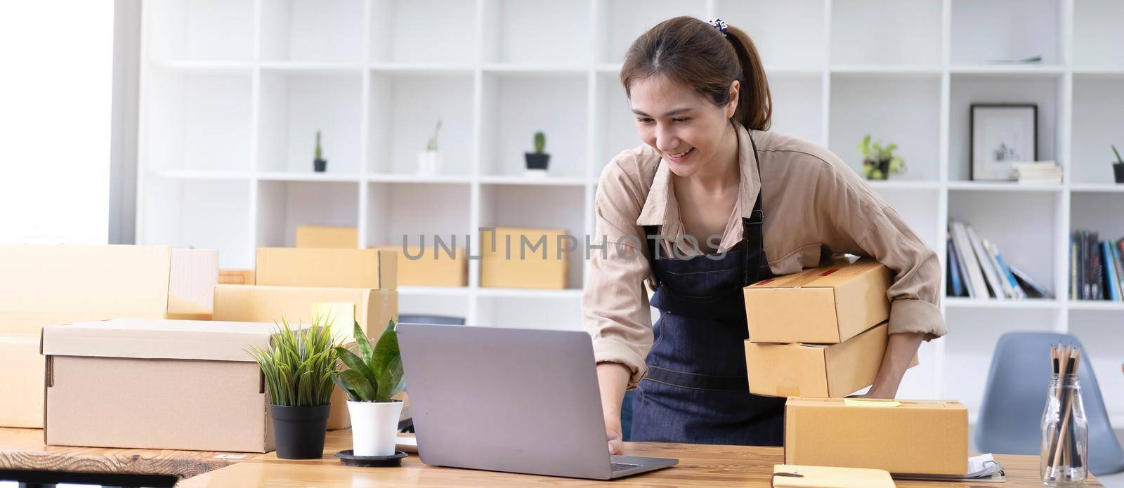 Young smiling beautiful owner asian woman freelancer sme business online shopping working on laptop computer with parcel box at home - SME business online and delivery concept by wichayada