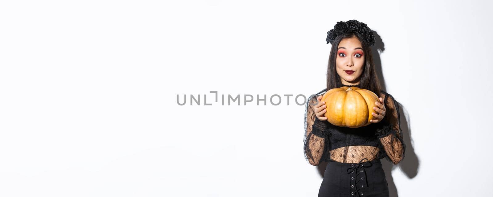 Portrait of excited pretty asian woman in halloween costume, dressed-up as witch or widow and holding pumpkin, standing over white background.
