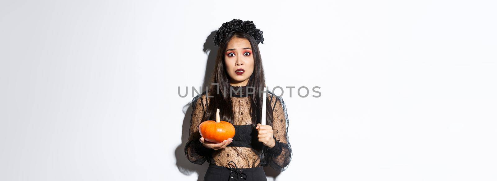 Image of asian woman panicking, being scared of witchcraft on halloween, standing in witch costume with candle and pumpkin by Benzoix