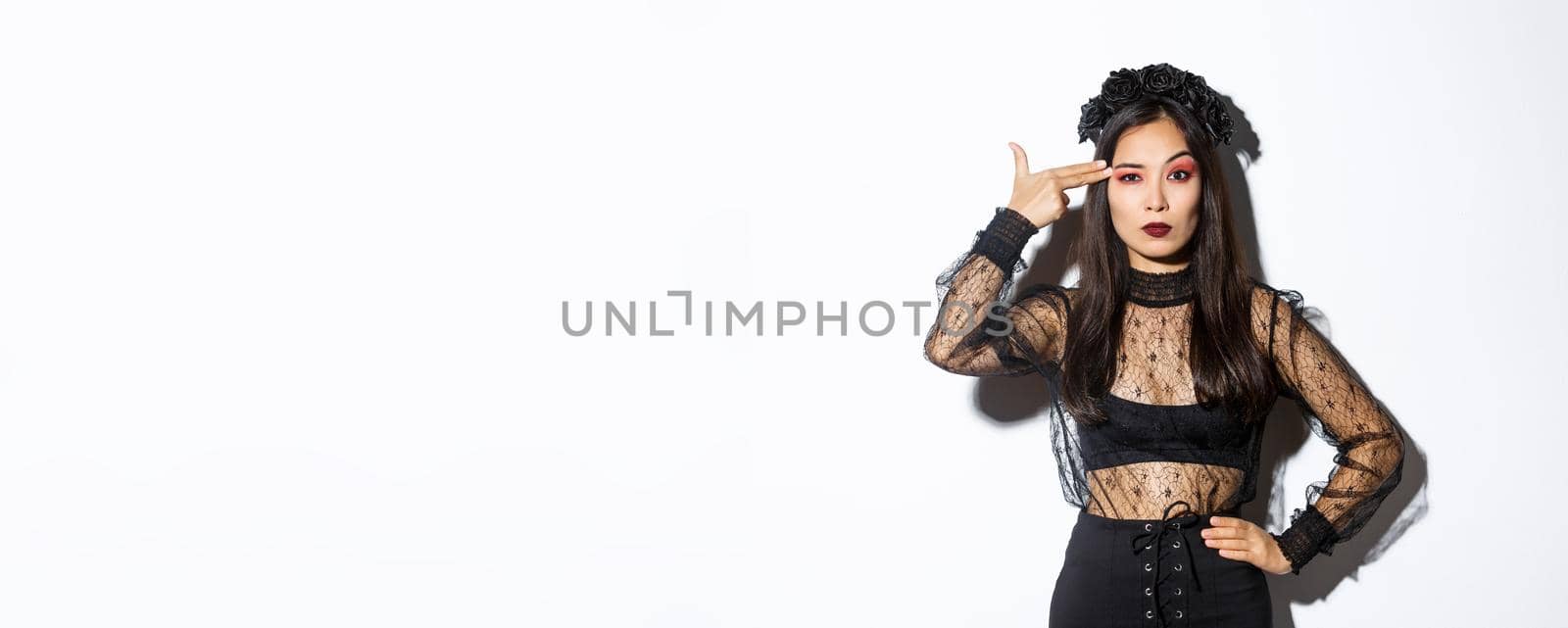 Annoyed young woman looking disappointed while making finger gun gesture over head, wearing halloween costume, standing over white background.