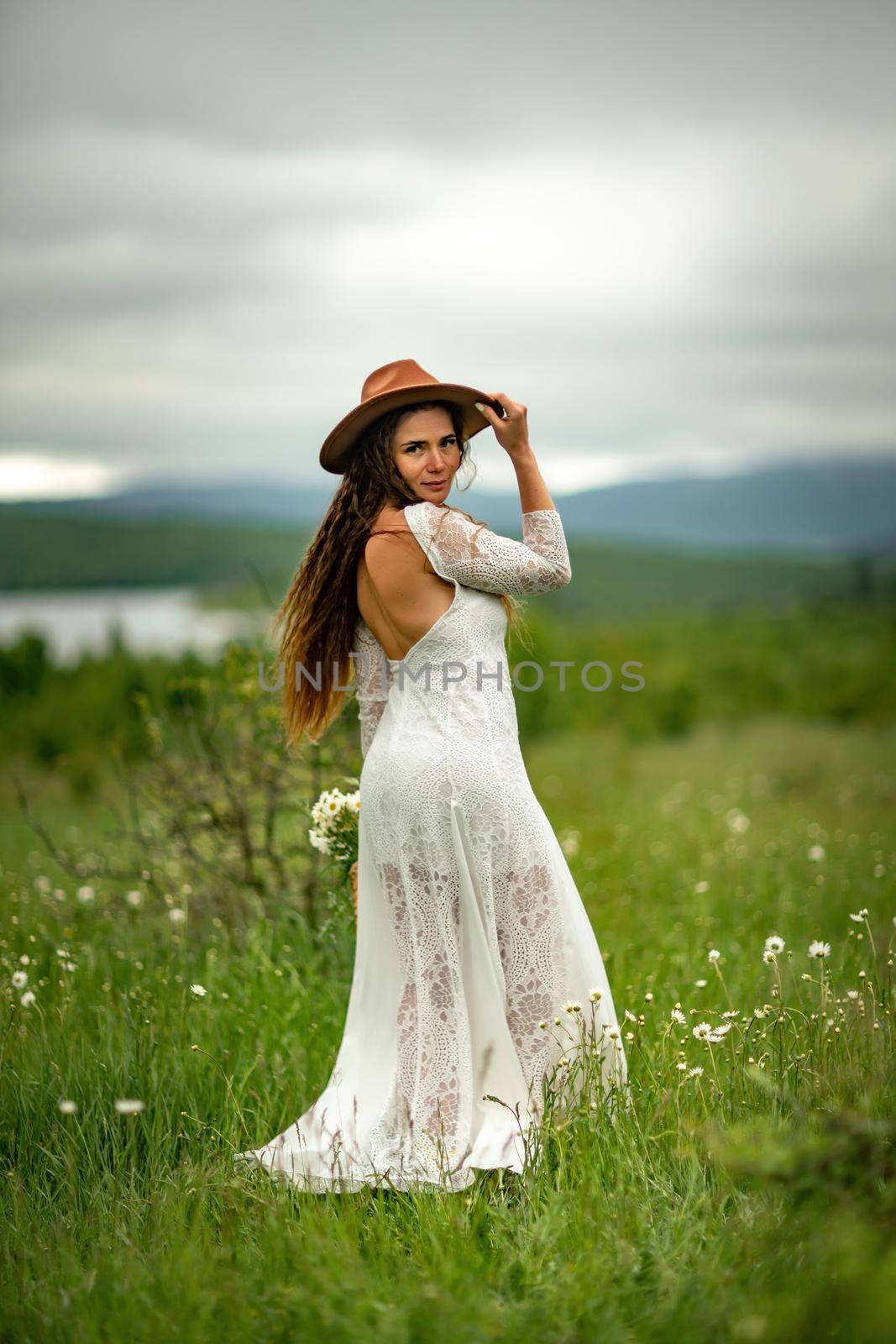 A middle-aged woman in a white dress and brown hat stands on a green field and holds a basket in her hands with a large bouquet of daisies. In the background there are mountains and a lake