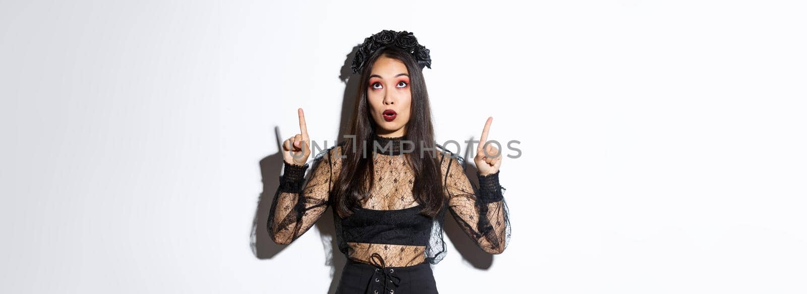 Surprised attractive asian woman in witch outfit pointing fingers up, showing halloween banner, promo of party. Beautiful female in black gothic dress looking amazed.