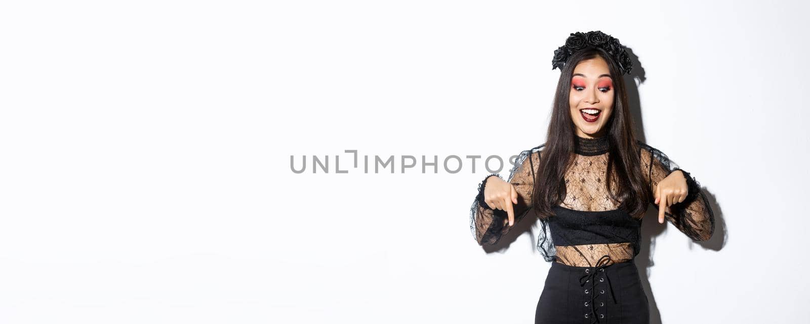 Excited asian woman dressed-up for halloween, smiling amused and pointing fingers down, showing advertisement, standing in party costume over white background by Benzoix