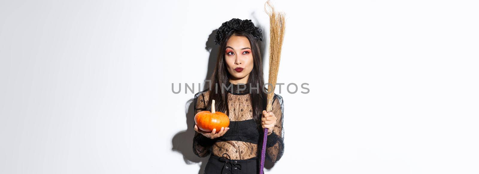 Image of sassy witch in gothic lace dress, holding broom and pumpkin, looking at upper left corner with halloween banner, white background by Benzoix