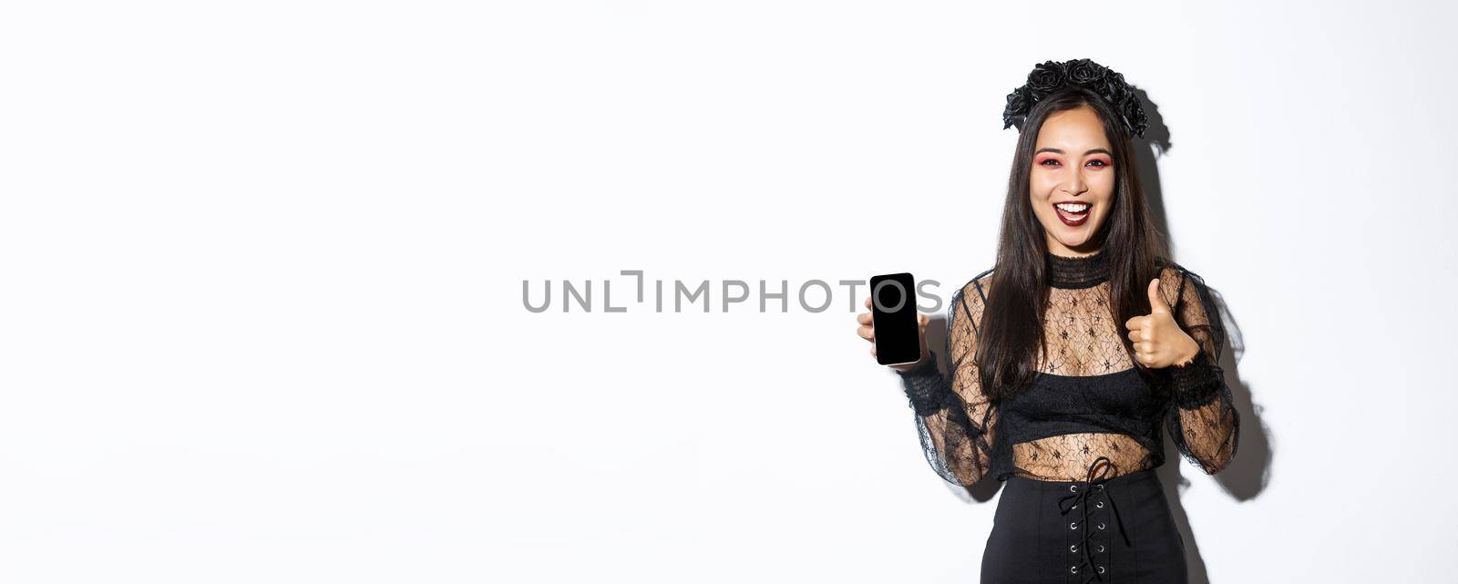 Excited and satisfied asian woman in halloween costume showing thumbs-up in approval and demonstrate mobile phone screen, standing over white background.