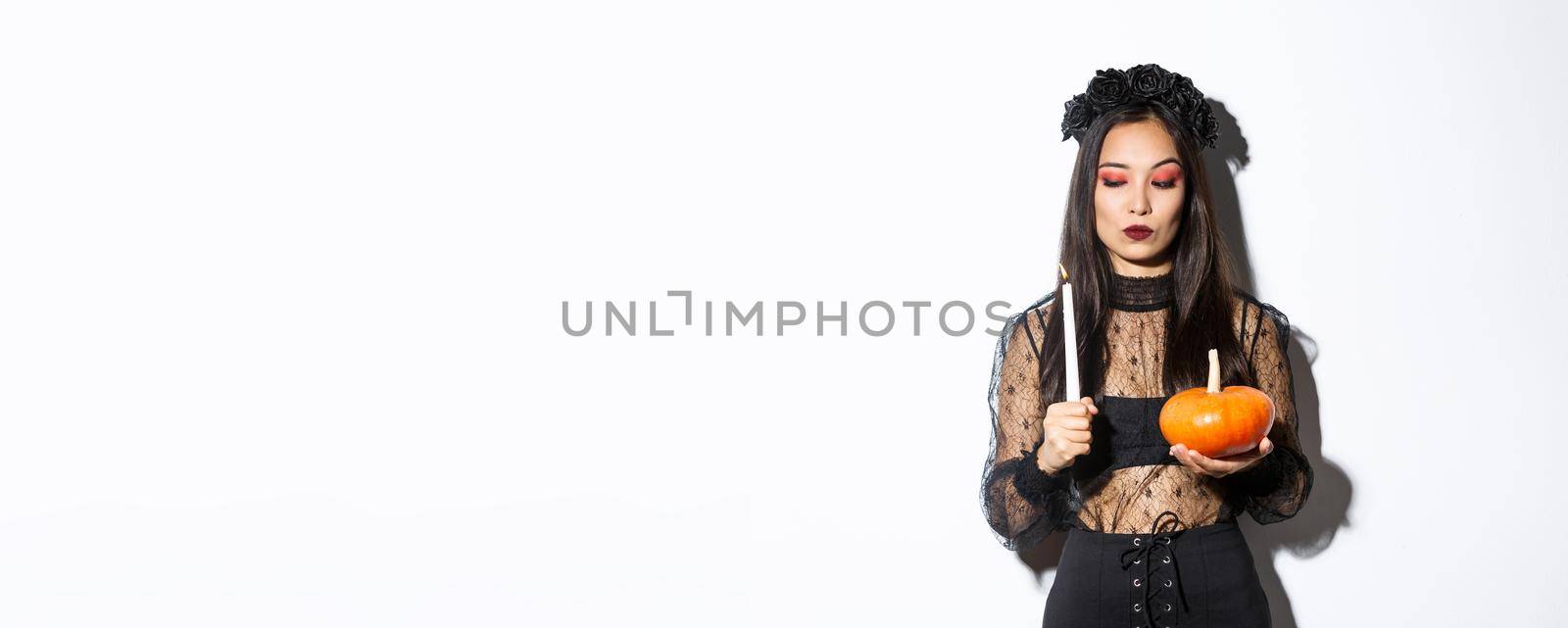 Myserious asian wicked witch in gothic dress, looking at lit candle, holding pumpkin, standing over white background.