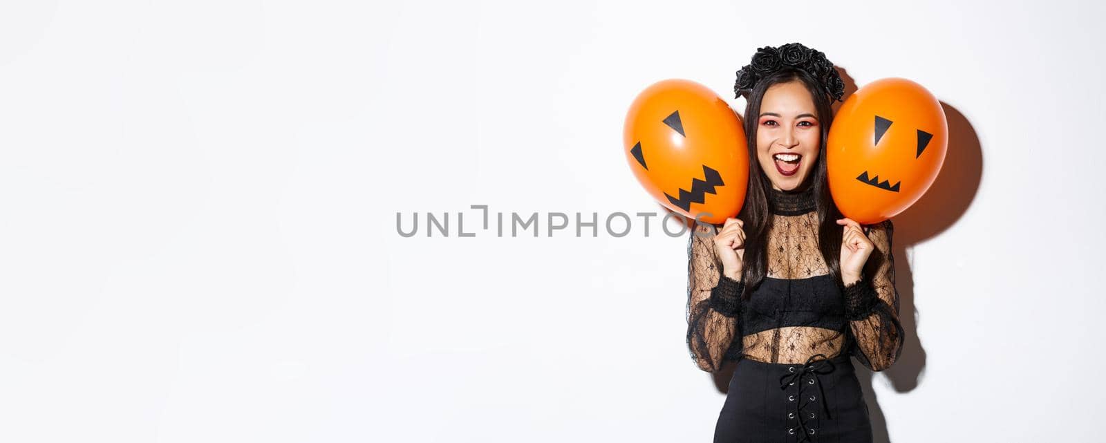 Image of asian girl in evil witch costume holding two orange balloons with scary faces, celebrating halloween, standing over white background by Benzoix