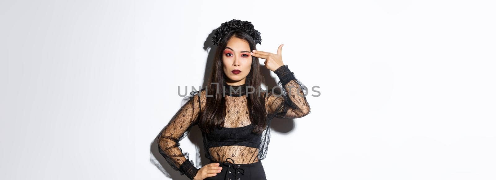Annoyed young woman looking disappointed while making finger gun gesture over head, wearing halloween costume, standing over white background.