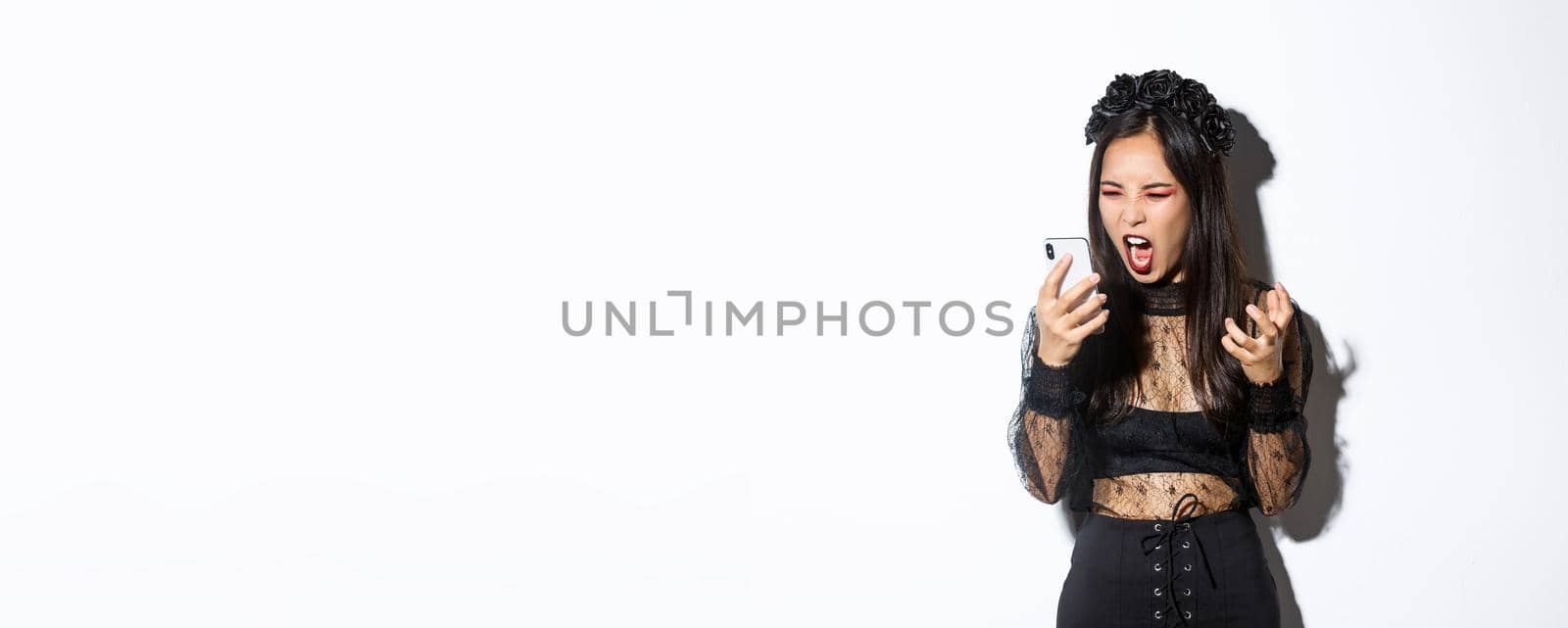 Portrait of angry asian woman in halloween costume looking mad, shouting at mobile phone and grimacing furious, standing over white background.
