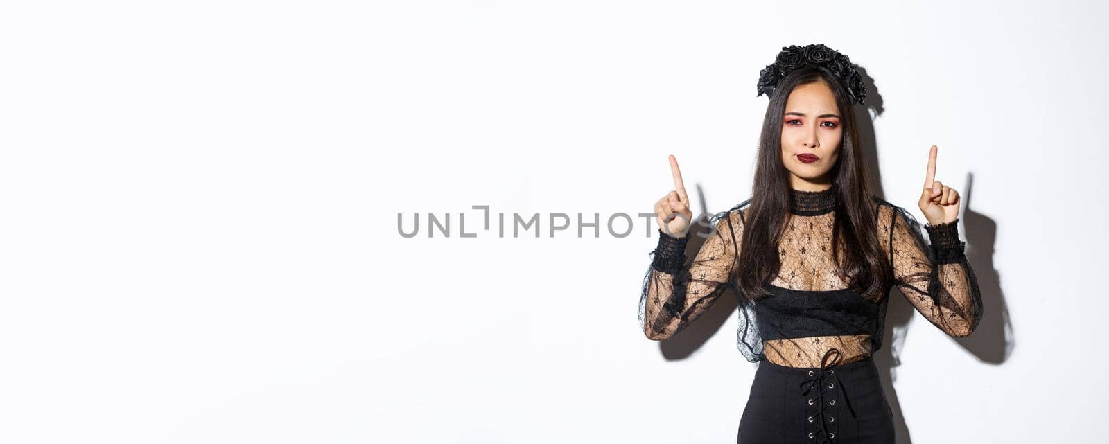 Disappointed and skeptical asian female in black dress and wreath looking displeased, pointing fingers up at something bad, have negative opinion, standing over white background by Benzoix