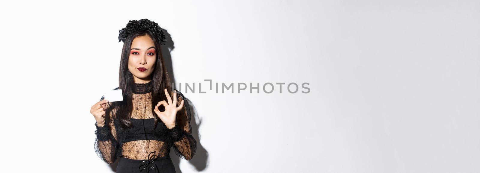 Portrait of confident asian woman assuring you in something, wearing halloween costume, showing okay gesture and credit card, white background.