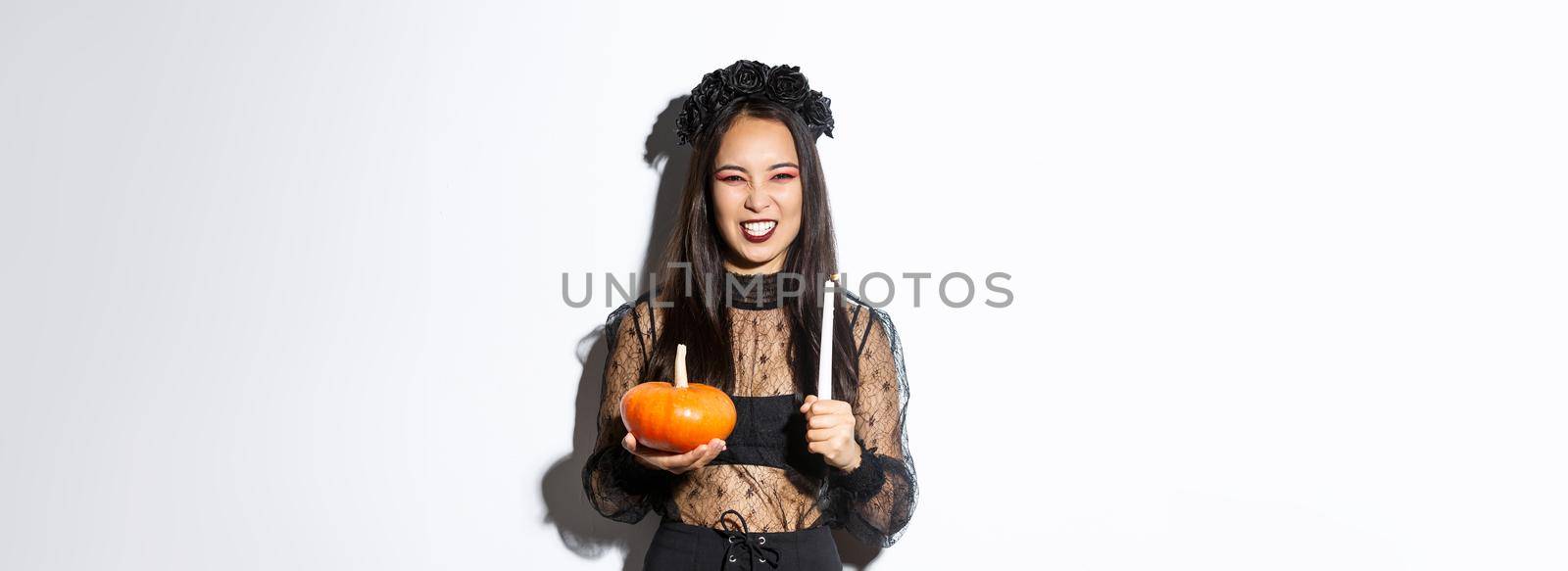 Image of asian evil witch in gothic lace dress and black wreath, laughing and grimacing, holding candle with pumpkin, celebrating halloween.