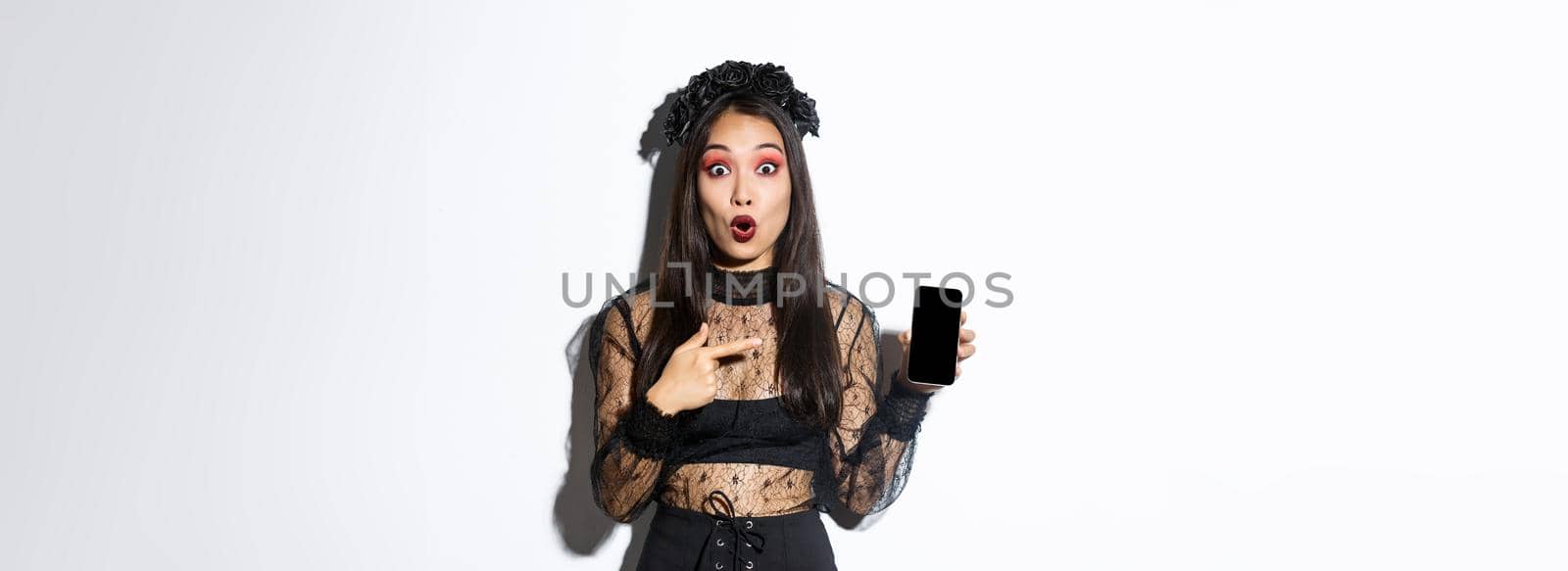 Surprised asian girl in black gothic dress with wreath, gasping amused and pointing finger at mobile phone display, showing halloween banner or promo, standing over white background.