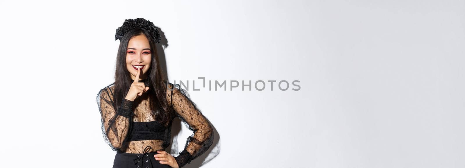 Smiling beautiful asian woman in black lace dress celebrating all saints day, have surprise for halloween, shushing with finger pressed to lips, share a secret, standing over white background by Benzoix
