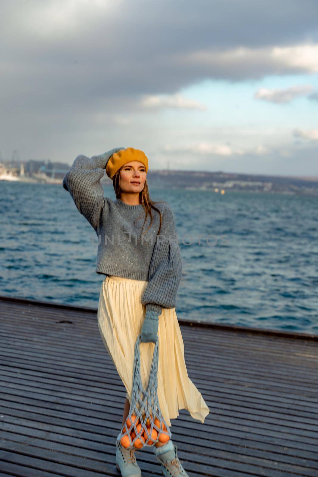 Outdoors fashion portrait of a beautiful middle aged woman walking on the beach. Marine background. Dressed in a stylish warm blue sweater, yellow skirt and beret