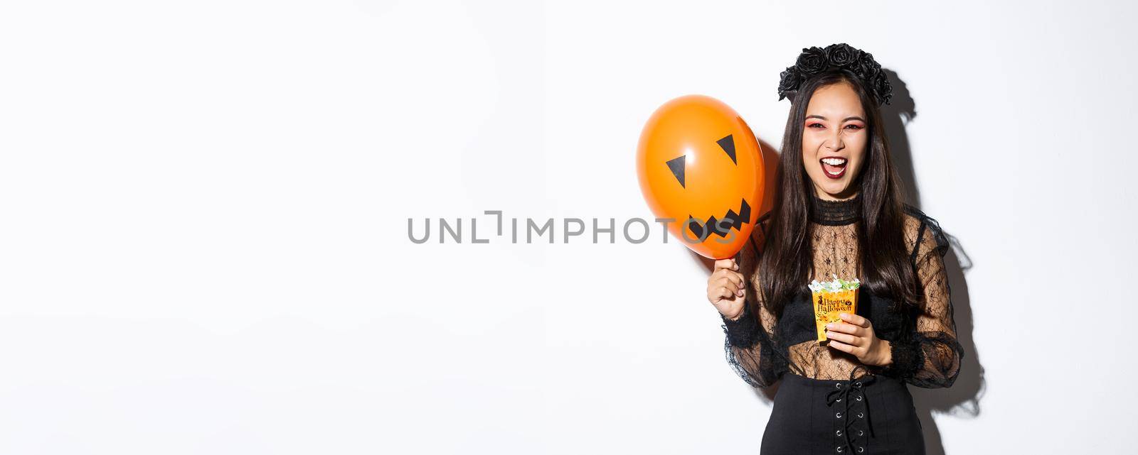 Scary witch trick or treating on halloween, holding sweets and orange balloon, standing in gothic lace dress with black wreath by Benzoix