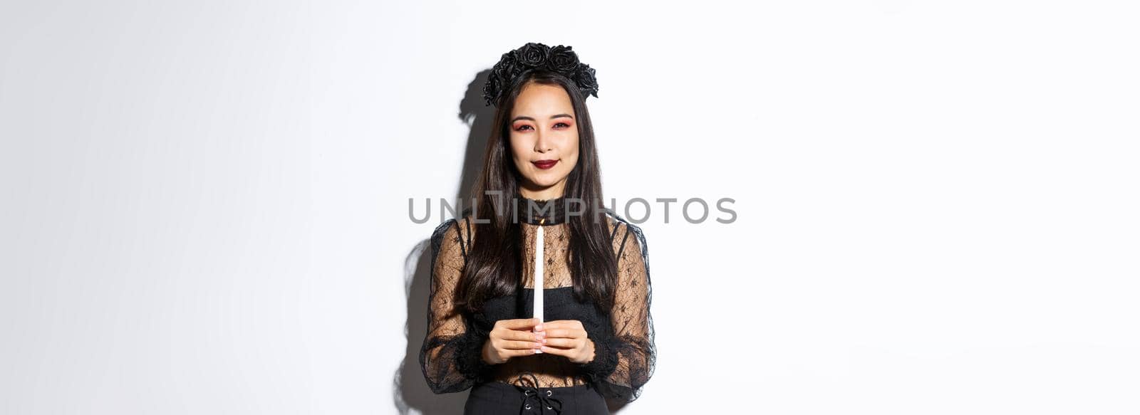 Image of beautiful woman celebrating halloween in witch costume, holding candle and squinting at camera suspicious, standing over white background.