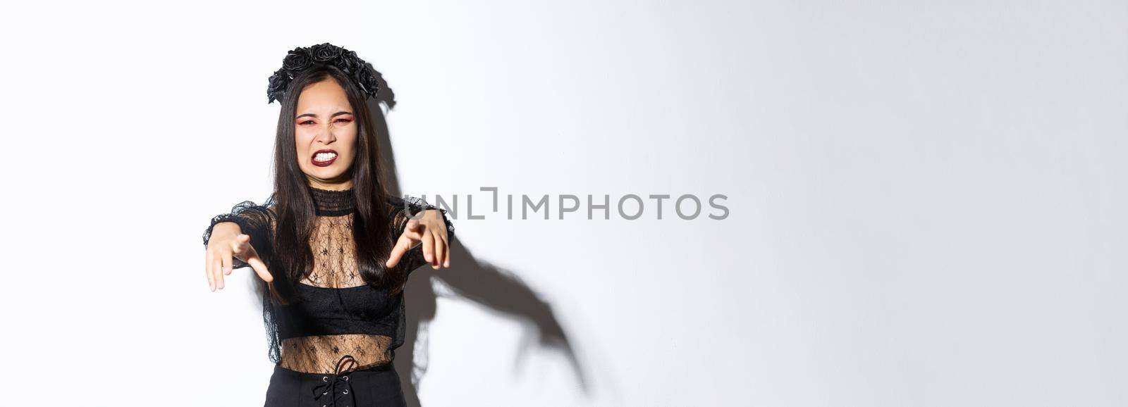 Image of beautiful undead evil witch cursing someone on halloween. Girl in party costume extend hands foward and looking scary, going for trick or treat, standing over white background.