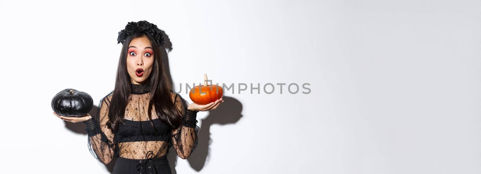 Portrait of surprised asian girl in witch costume holding two pumpkins and gasping amazed while looking at camera, standing over white background by Benzoix