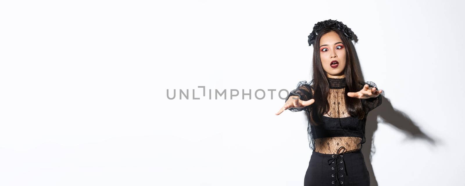 Image of funny and beautiful woman acting like undead widow, wearing black lace dress and wreat, looking like zombie and moving forward. Female enjoying halloween party.