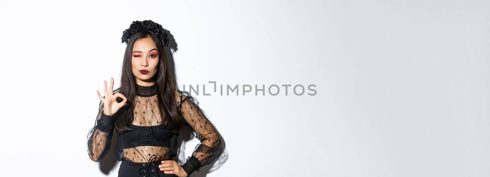 Sassy beautiful asian woman in witch costume showing okay gesture, all under control. Confident girl in halloween gothic dress like or approve something, standing white background by Benzoix