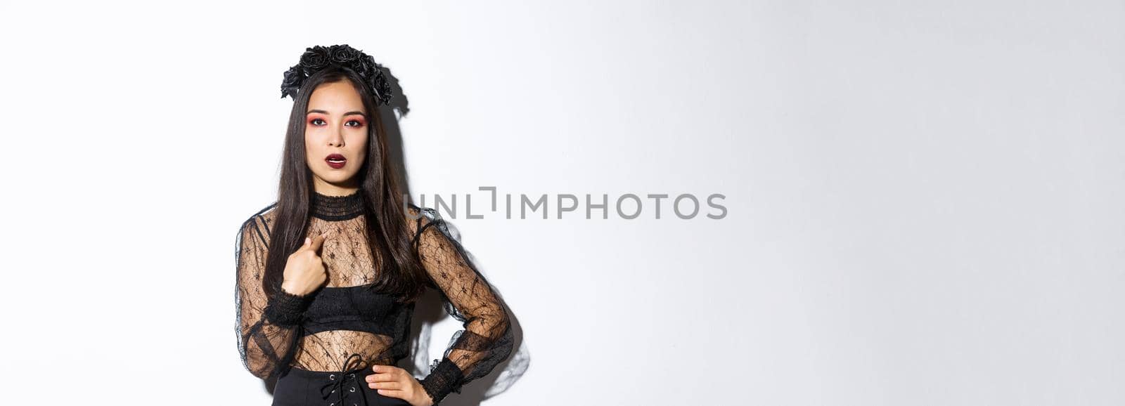 Annoyed and confused asian woman in witch halloween costume pointing at herself, looking bothered and unamused, standing reluctant over white background.