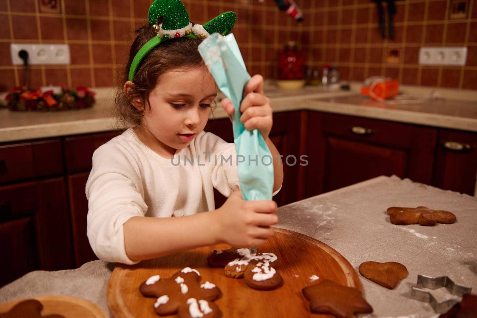 Beautiful child, a lovely Caucasian little girl in white sweater and elf hoop, squeezing icing from a baking bag on baked gingerbread cookies. Child learns decorating pastries with glaze for Christmas