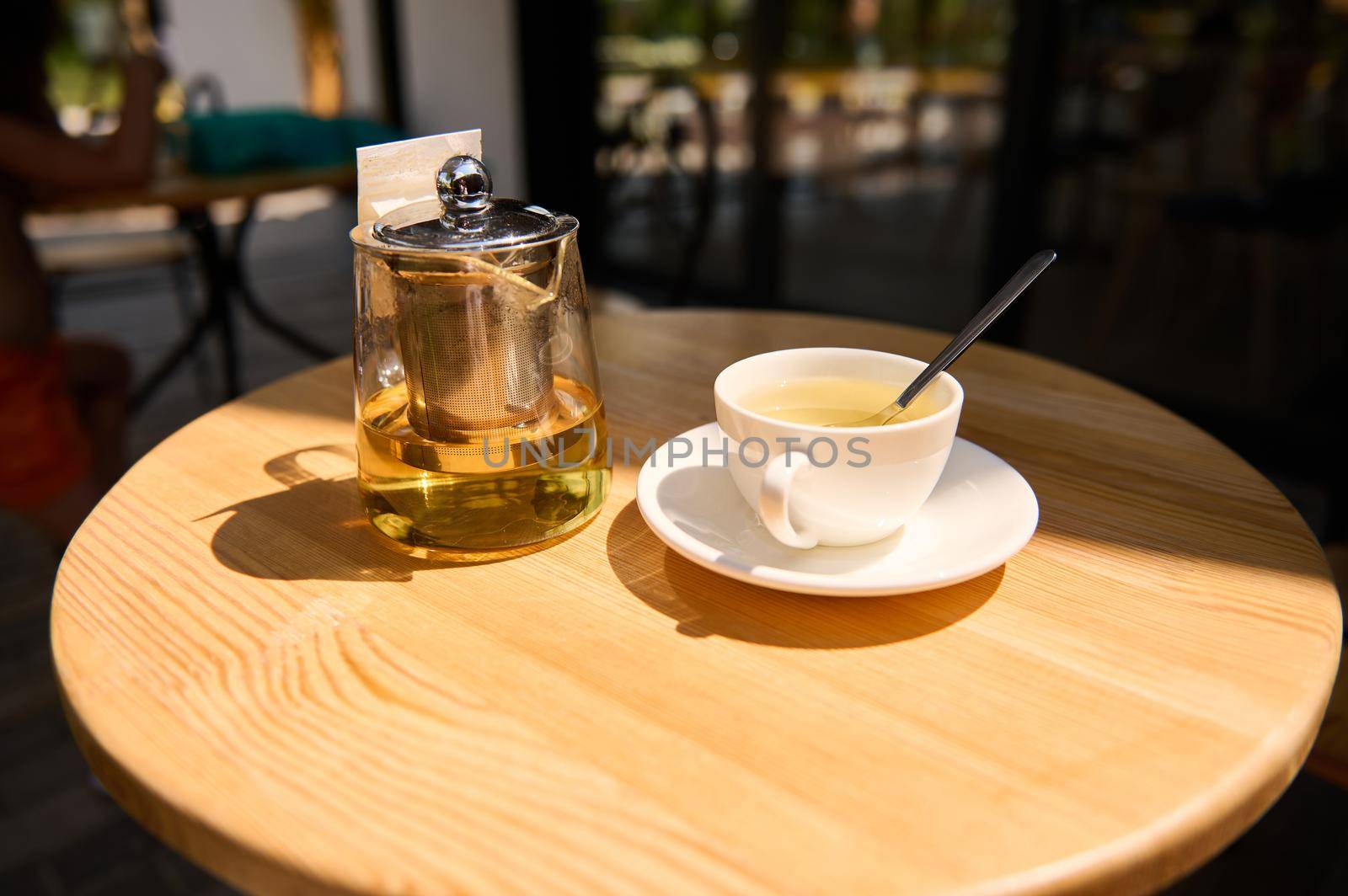 A teacup on a white saucer and glass teapot with healthy healing and antioxidant herbal tea, on a wooden round table in the cafeteria summer terrace. Antioxidant healthy and healing hot drink. Detox