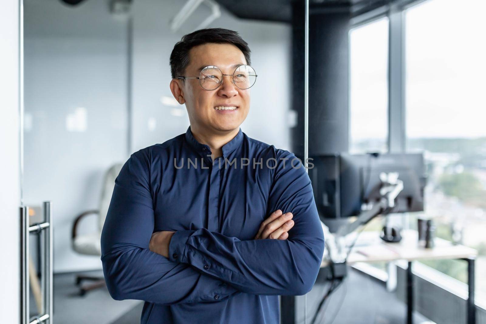 Portrait of Asian businessman, man in glasses and shirt looking out window and smiling, worker inside office building with crossed arms by voronaman