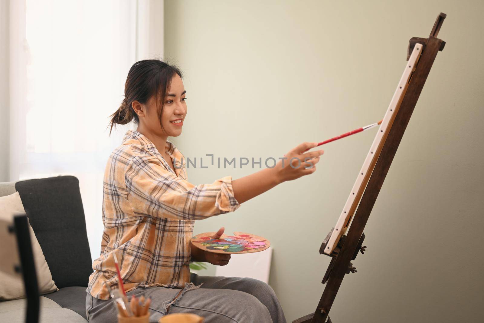 Satisfied female painter painting on easel with watercolor in comfortable workshop. Leisure activity, creative hobby and art concept.