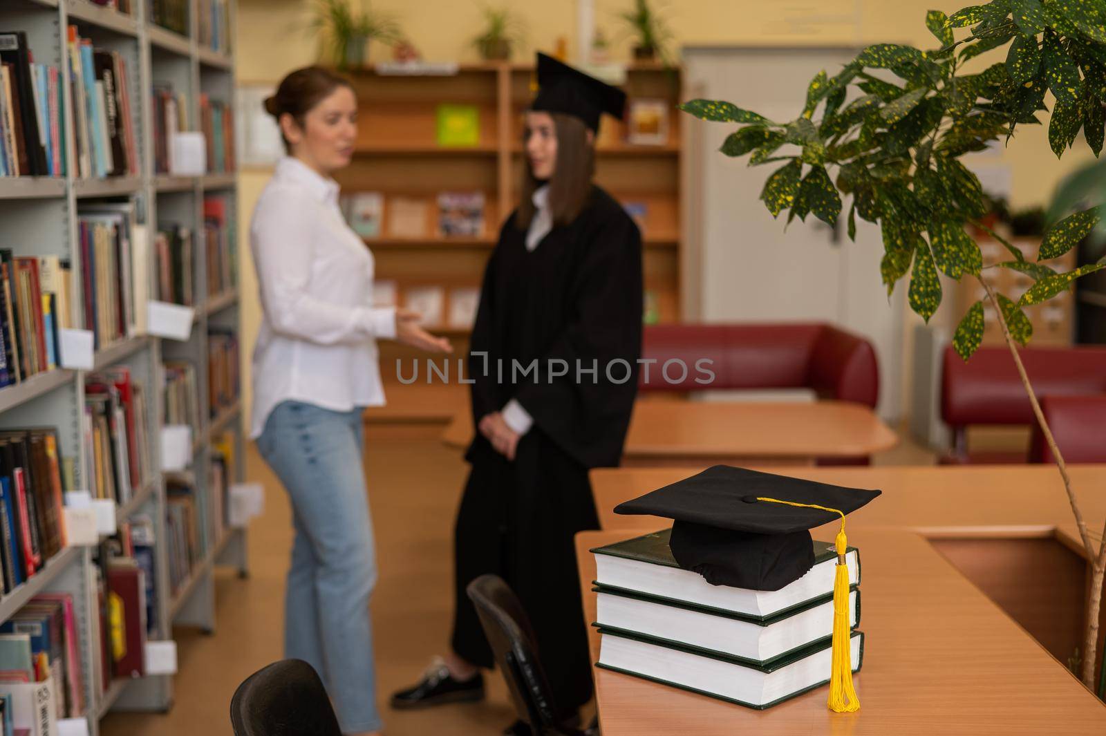 Close-up of a graduation cap on a stack of books in a library. teacher and young female graduate in the background. by mrwed54