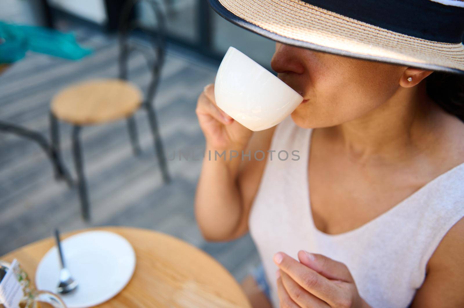 Close-up of a beautiful mysterious Latin American woman, in a straw hat covering half of her face, and elegant summer dress, sitting at a table in a summer terrace, enjoying her coffee break outdoors