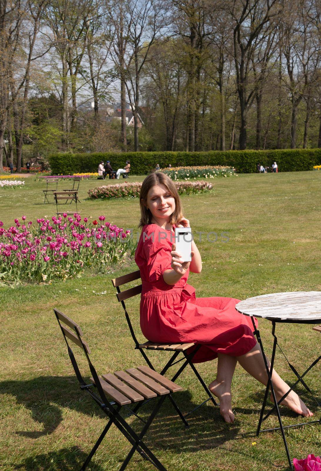 A young beautiful girl in a red dress in a spring park plays with her hair, flirts and takes a selfie on a mobile phone, tulips bloom and people walk in the park, pamper themselves.High quality photo