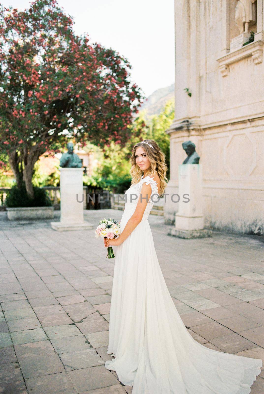 Bride in a white dress with a bouquet stands near an ancient building with sculptures. High quality photo